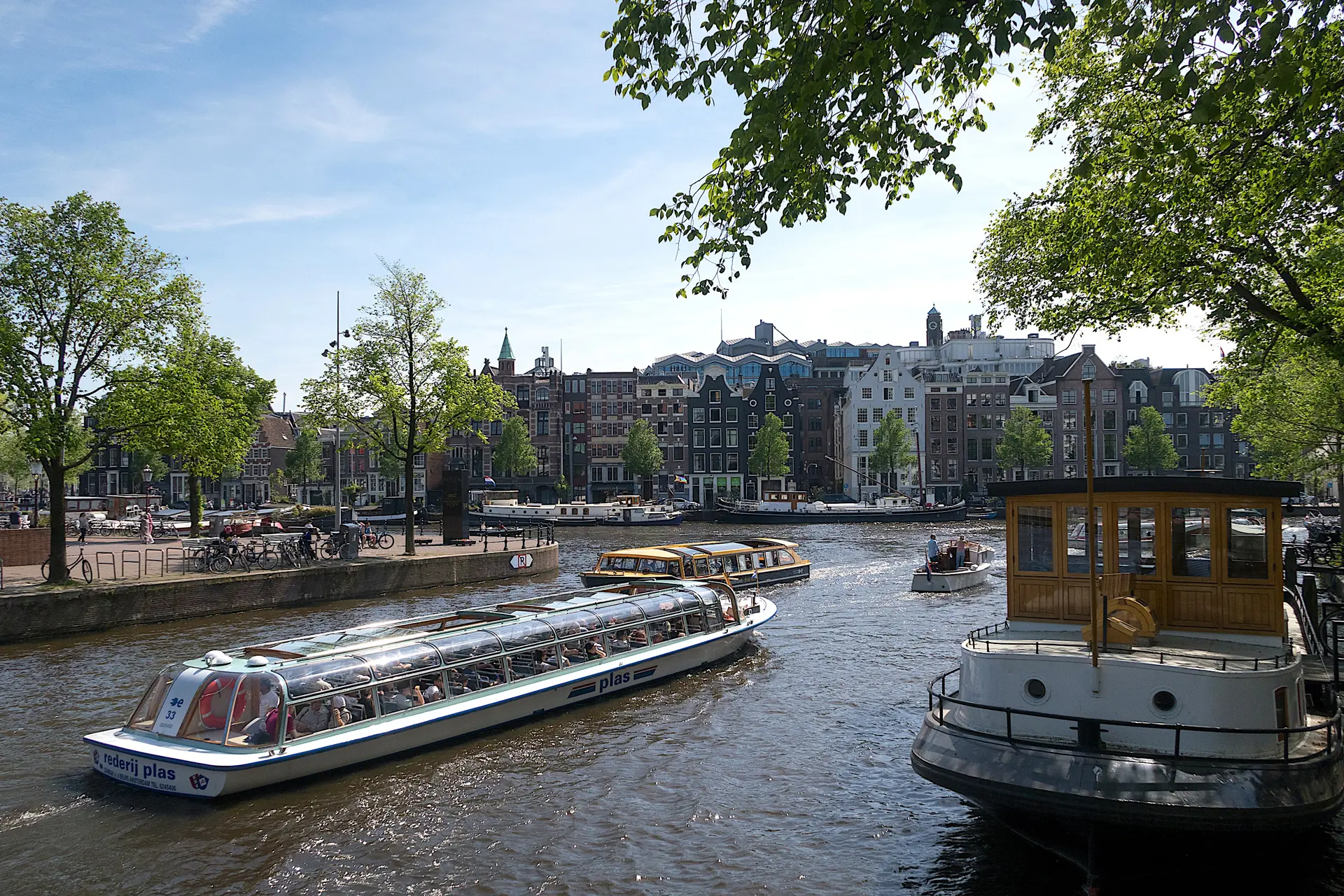 Les bateaux touristiques naviguent sur un canal d'Amsterdam bordé d'arbres et de bâtiments historiques, tandis que les visiteurs admirent le paysage — l'idéal pour ceux qui souhaitent passer la nuit à Amsterdam sous un ciel bleu et dégagé.