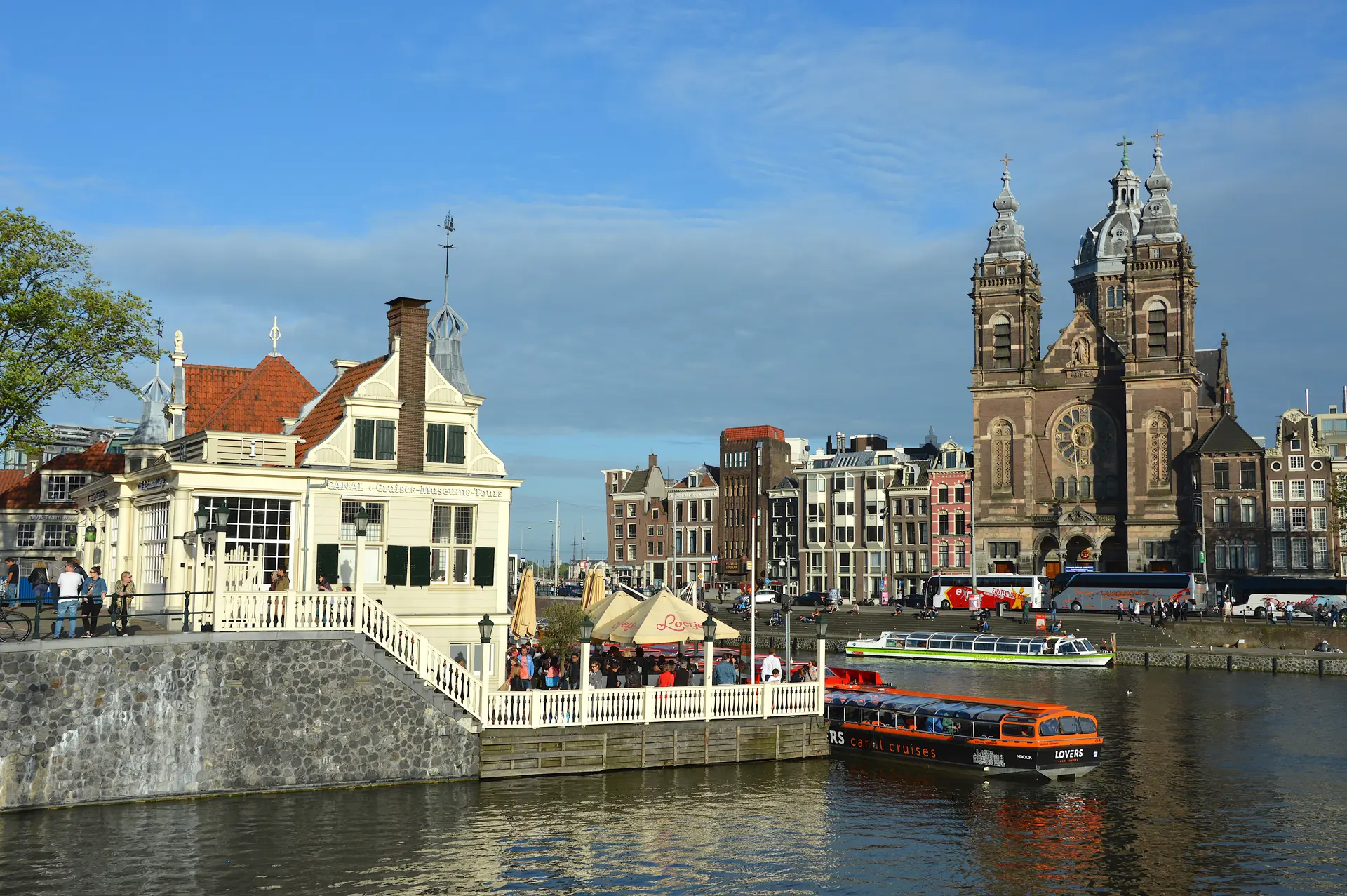 Un canal d'Amsterdam avec un bateau-mouche, des bâtiments historiques, une terrasse de café et les tours jumelles de la basilique Saint-Nicolas sous un ciel bleu : un incontournable pour tous ceux qui souhaitent visiter Amsterdam.