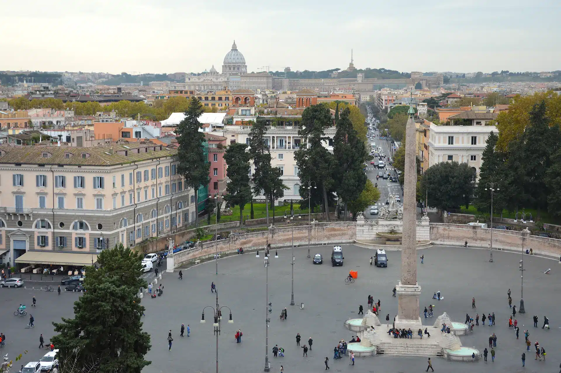 Vue de la Piazza del Popolo à Rome, avec un obélisque au centre, entouré de personnes et de bâtiments historiques, la basilique Saint-Pierre étant visible à l'arrière-plan.