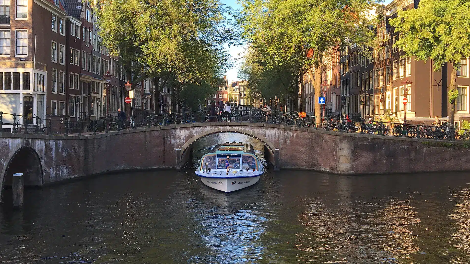 Un bateau-mouche blanc passe sous un pont en briques sur un canal bordé de grands arbres et de bâtiments historiques à Amsterdam ; on aperçoit des passants et des vélos sur le pont.