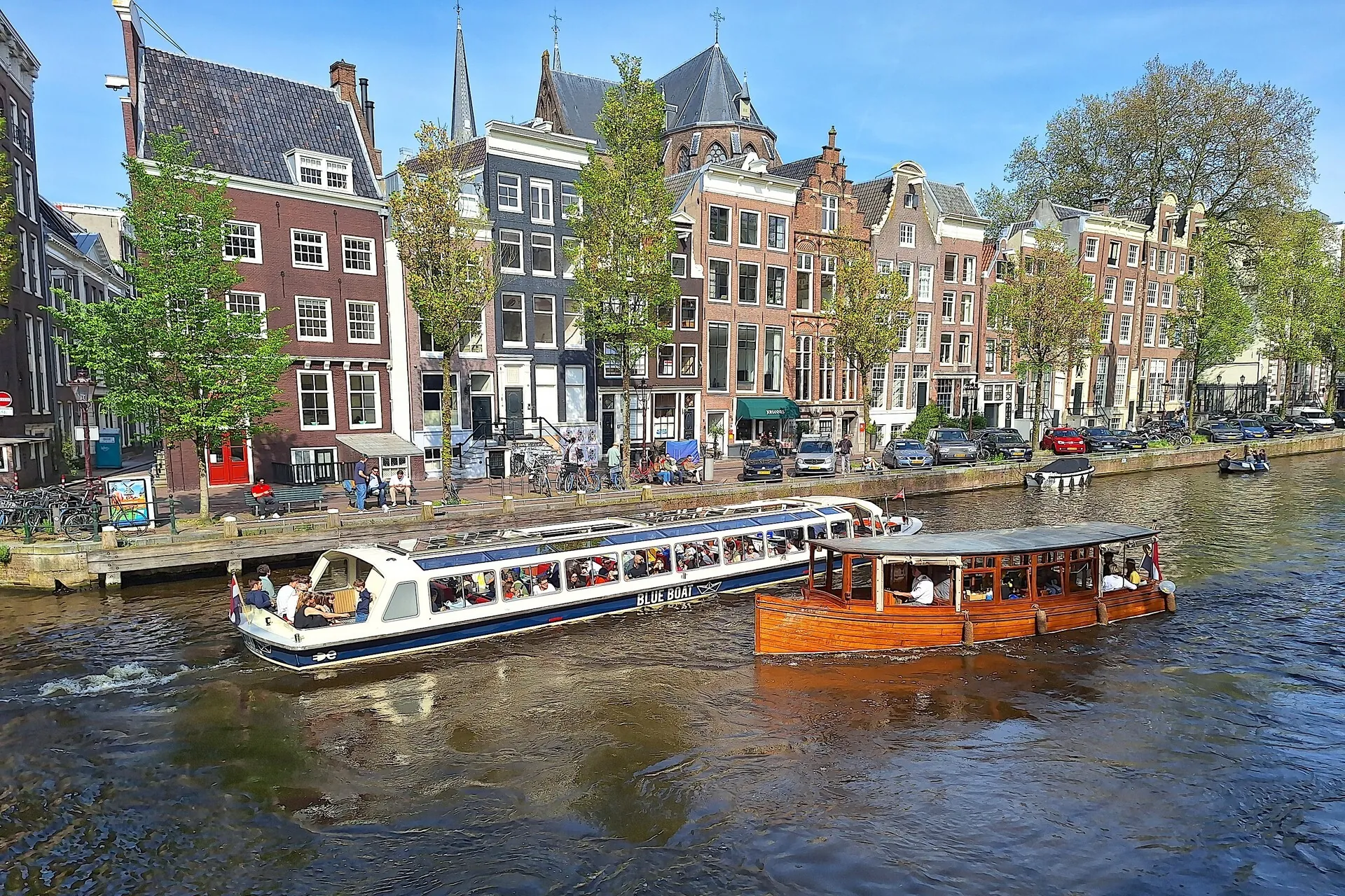 Par une journée ensoleillée à Amsterdam, deux bateaux transportant des passagers glissent sur un canal, longeant des bâtiments traditionnels néerlandais et des arbres. Des voitures garées bordent la rue, contribuant au charme de la ville pour ceux qui souhaitent visiter Amsterdam en trois jours.