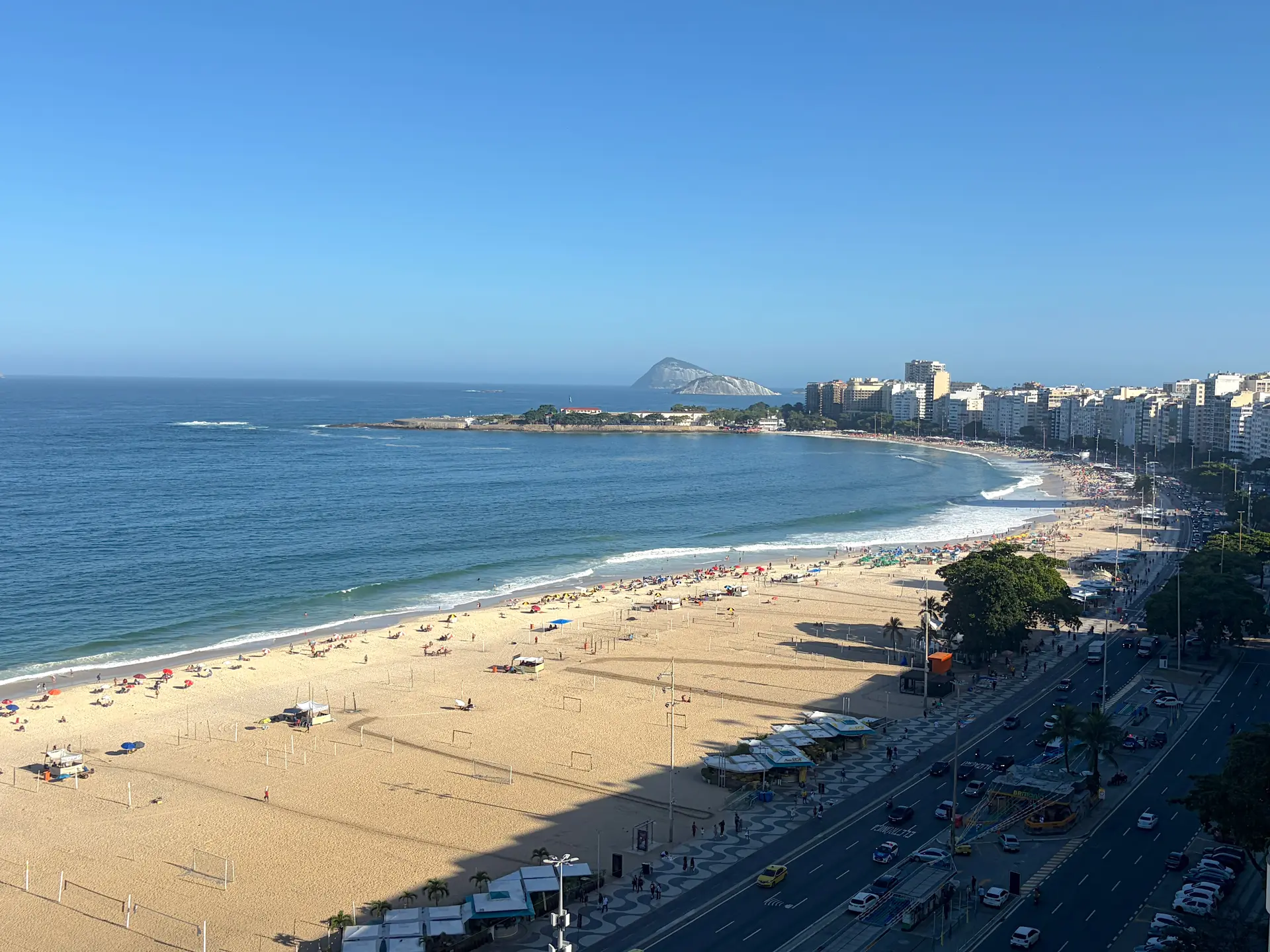 Vue panoramique d'une plage ensoleillée avec des gens sur le sable, un océan bleu, des immeubles en bord de mer et une route bordée d'arbres sur laquelle circulent des voitures. Une colline lointaine se profile à l'horizon sous un ciel bleu limpide.