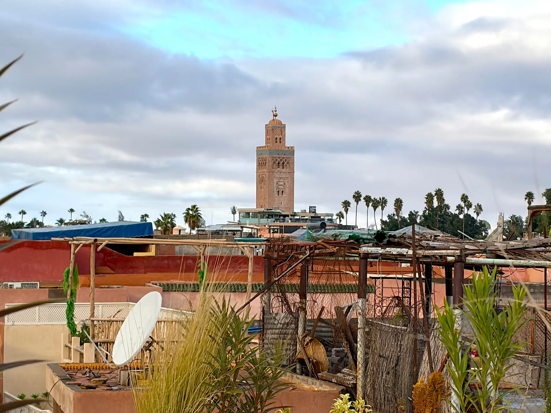 Une vue depuis un toit, avec des plantes et une antenne parabolique au premier plan, et la tour de la mosquée Koutoubia se dressant à l'arrière-plan sous un ciel nuageux à Marrakech, au Maroc : une vue idéale pour ceux qui souhaitent visiter la médina de Marrakech et s'imprégner de son atmosphère animée.